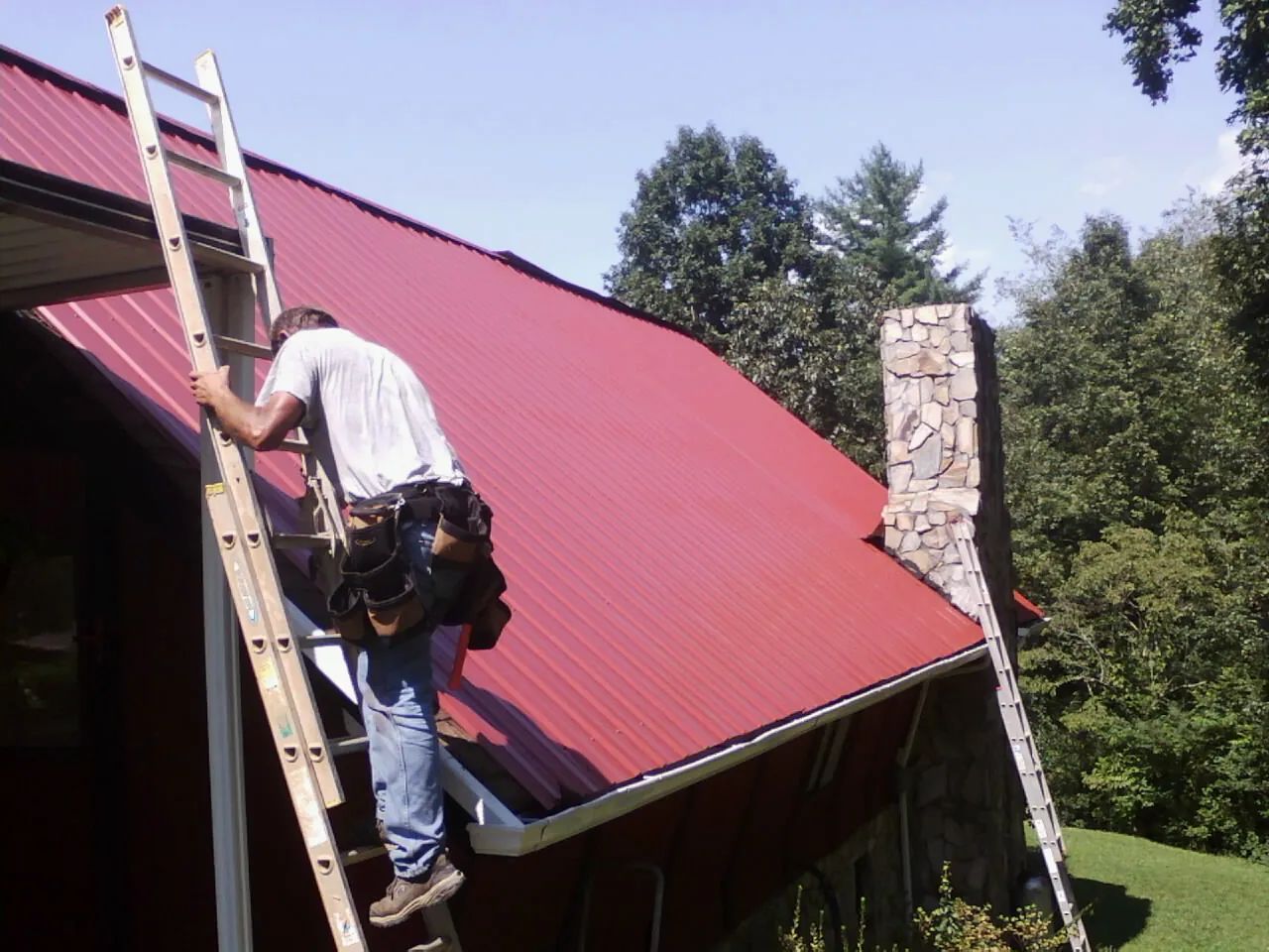 Roofer on a ladder beside a red metal roof and stone chimney — Soffit & Fascia Repair by Roofing Brothers in Alamance